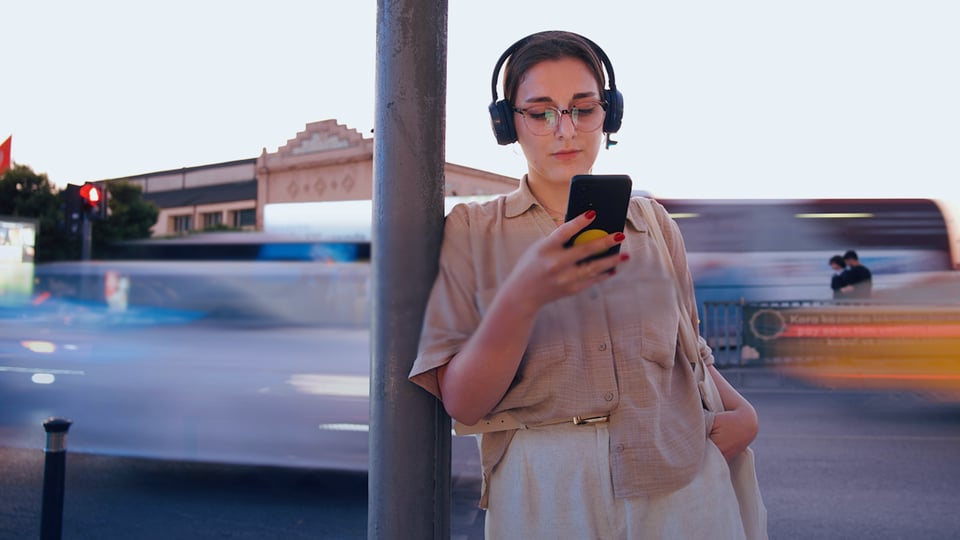 En ung person står vid en stolpe i stadsmiljö med hörlurar och tittar på sin mobiltelefon. I bakgrunden syns förbipasserande fordon i rörelse som ger en suddig effekt.